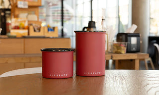 Airscape Kaffeedosen in Ziegelrot, Medium und Small, auf Holztisch im Café – Edelstahlbehälter für frische Kaffeebohnen und Gewürze