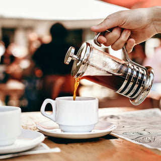 Frisch gebrühter Kaffee wird aus einer French Press in eine weiße Tasse auf einem Café-Tisch gegossen.