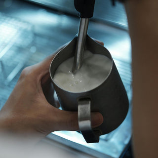 Milch wird in einem Edelstahlkännchen mit der Dampfdüse einer Espressomaschine aufgeschäumt.