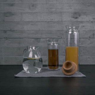 Drei Glaskrüge von Carl Henkel, darunter der MIZU Krug, mit Wasser, Tee und Saft auf einem dunklen Tisch vor Betonwand.