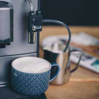 Nahaufnahme einer NIVONA CafeRomatica mit Milchdüse, silbernem Milchkännchen im Hintergrund und türkisfarbener Kaffeetasse mit Milchschaum auf der Abtropfschale.