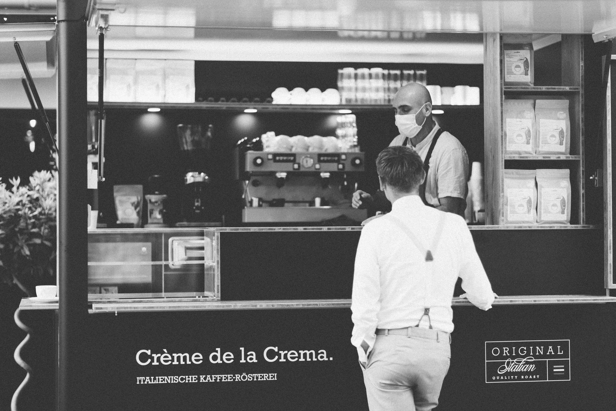 Schwarz-weißes Foto von Roberto Cascone beim Bedienen eines Kunden am Cremaexpress-Coffeetruck. Neben ihm steht eine Espressomaschine und im Hintergrund sind Tassen und AMORI-Kaffeetüten zu sehen. Ein Kunde gibt eine Bestellung auf. Das Bild könnte während einer Hochzeit oder eines Events aufgenommen worden sein.