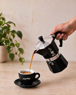Eine Hand gießt frisch gebrühten Kaffee aus einer schwarzen Bialetti Espressokanne in eine AMORI Tasse.