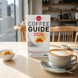 Crema Coffee Guide 2026 liegt auf einem Holztisch in einem Café neben einer Tasse Cappuccino