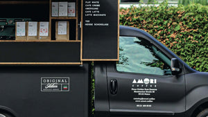 Ein Ausschnitt des Amori Coffee Trucks: Die seitliche Tür zeigt das Amori-Logo und Kontaktdaten. Links davon befindet sich der Verkaufsbereich mit verschiedenen Verpackungen von Amori Coffee und einer Tafel mit den angebotenen Kaffeegetränken.