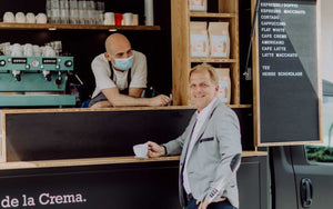 Ein Bild von Roberto Cascone, dem Inhaber von Amori Coffee, und einem Kunden, der vor dem Coffee-Truck Cremaexpress steht. Der Kunde hält eine Tasse Kaffee in der Hand, während Roberto lächelnd in Richtung Kunde schaut.