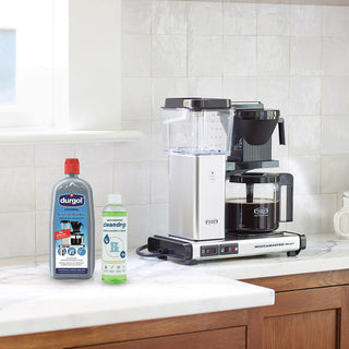 Durgol Universal Entkalker und Moccamaster Cleandrip nebeneinander auf Küchenarbeitsplatte, im Hintergrund Moccamaster Filterkaffeemaschine