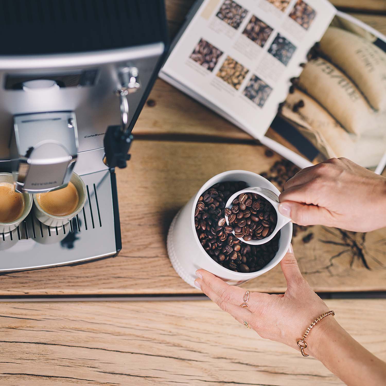 Person bereitet Kaffee mit einem NIVONA Vollautomaten zu. Zwei frisch zubereitete Espresso sind auf der Abtropfschale zu sehen, während die Person Kaffeebohnen aus einem Behälter in einen Messbecher schöpft. Im Hintergrund ist ein Kaffeebuch mit Kaffeebohnen und einem Kaffeesack abgebildet.