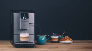 Auf dem Bild ist ein NIVONA Kaffeevollautomat im Edelstahl-Look (CafeRomatica NICR 795) zu sehen. Auf der Abtropfschale steht eine weiße Tasse gefüllt mit einem Milchkaffee. Rechts neben der Maschine befindet sich eine kleine grüne Milchkanne. Das Kännchen ist mit dem Kaffeevollautomaten per Milchschlauch verbunden.