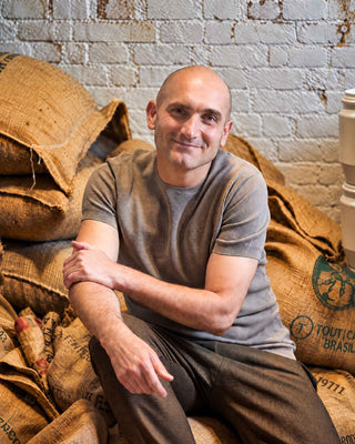 Roberto Cascone sitzt lächelnd mit verschränkten Armen auf gestapelten Kaffeesäcken vor einer weißen Backsteinwand.