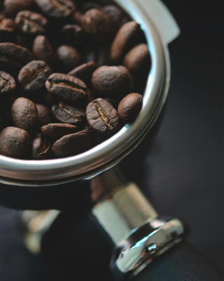 Nahaufnahme dunkelbraun gerösteter Kaffeebohnen im Metallsieb eines Siebträgers mit schwarzem Griff.