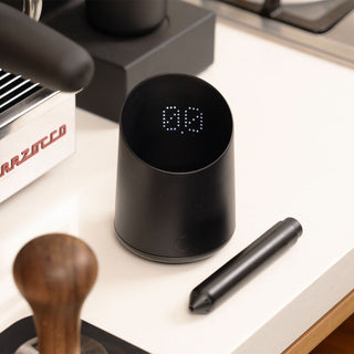 Subminimal Subscale Digitalwaage auf einer Espresso-Arbeitsfläche neben La Marzocco Maschine und Zubehör – LED-Anzeige aktiv, Kaffeestation im Hintergrund.
