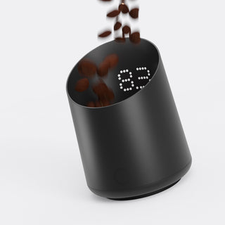 Schwarze Subminimal Subscale Digitalwaage in schräger Position, mit fallenden Kaffeebohnen und sichtbarer LED-Gewichtsanzeige.