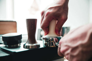 Die Hand eines Baristas presst mit einem weißen Tamper das Kaffeemehl in den Siebträger. Ein weiterer Tamper mit Holzgriff steht daneben.